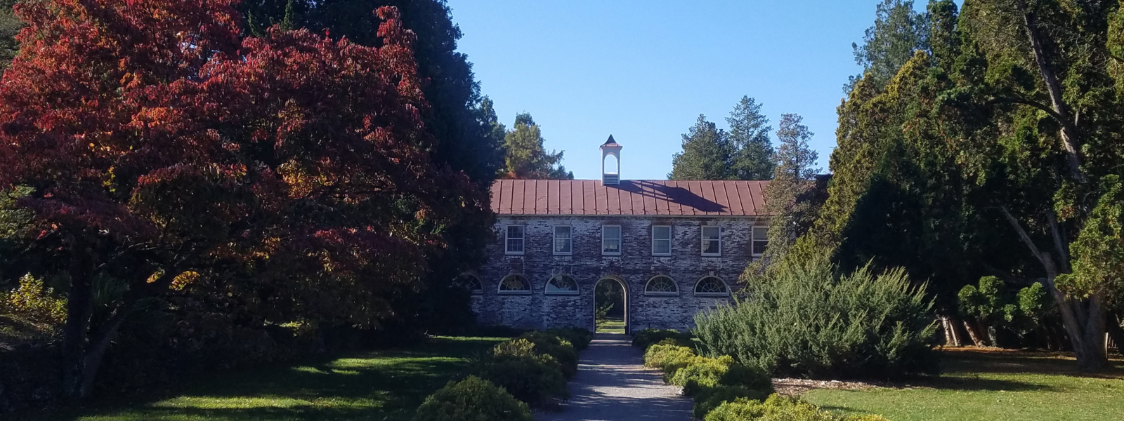Blandy's central quarters building, a brick structure with a red roof and open central archway, surrounded my conifers with a boxwood lined path leading up to it